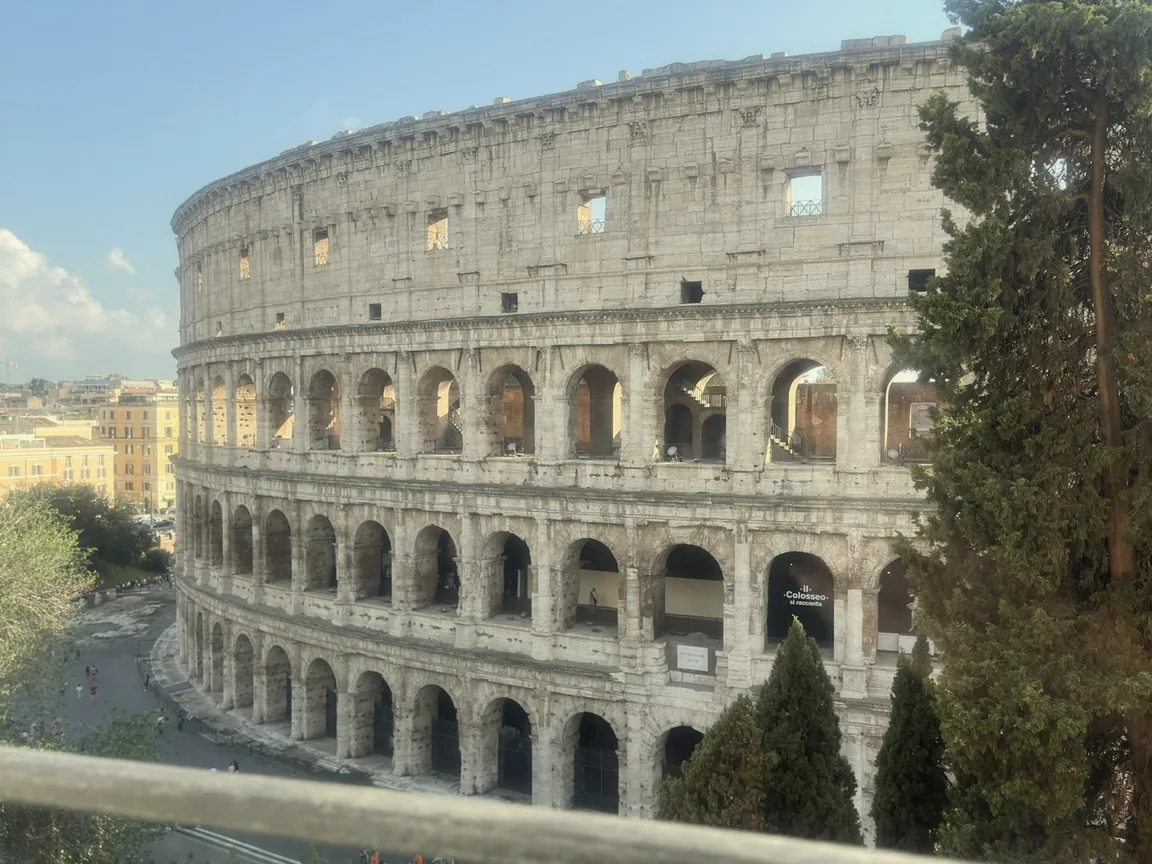 The Colosseum framed by cypress trees and a balcony railing under a bright blue Roman sky