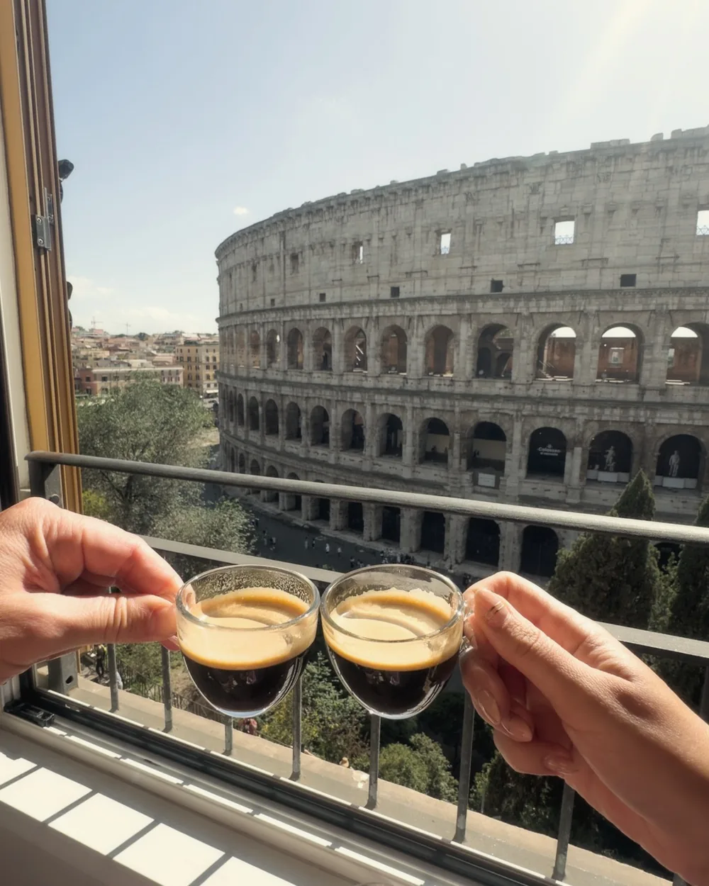Two hands clinking small glasses of espresso on the balcony, the Colosseum behind