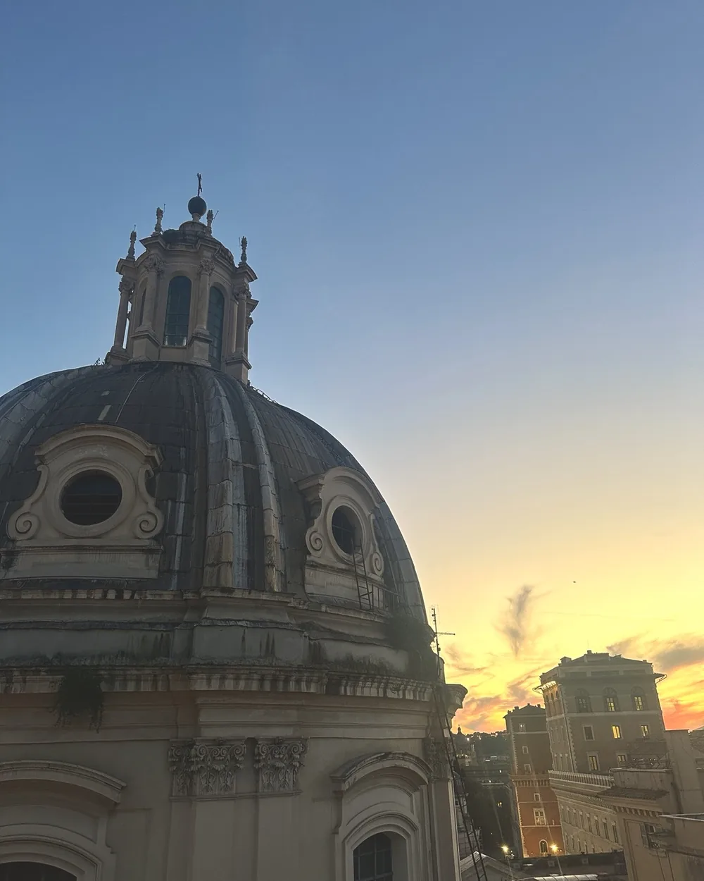 A baroque church dome silhouetted against a golden sunset sky over Rome's rooftops