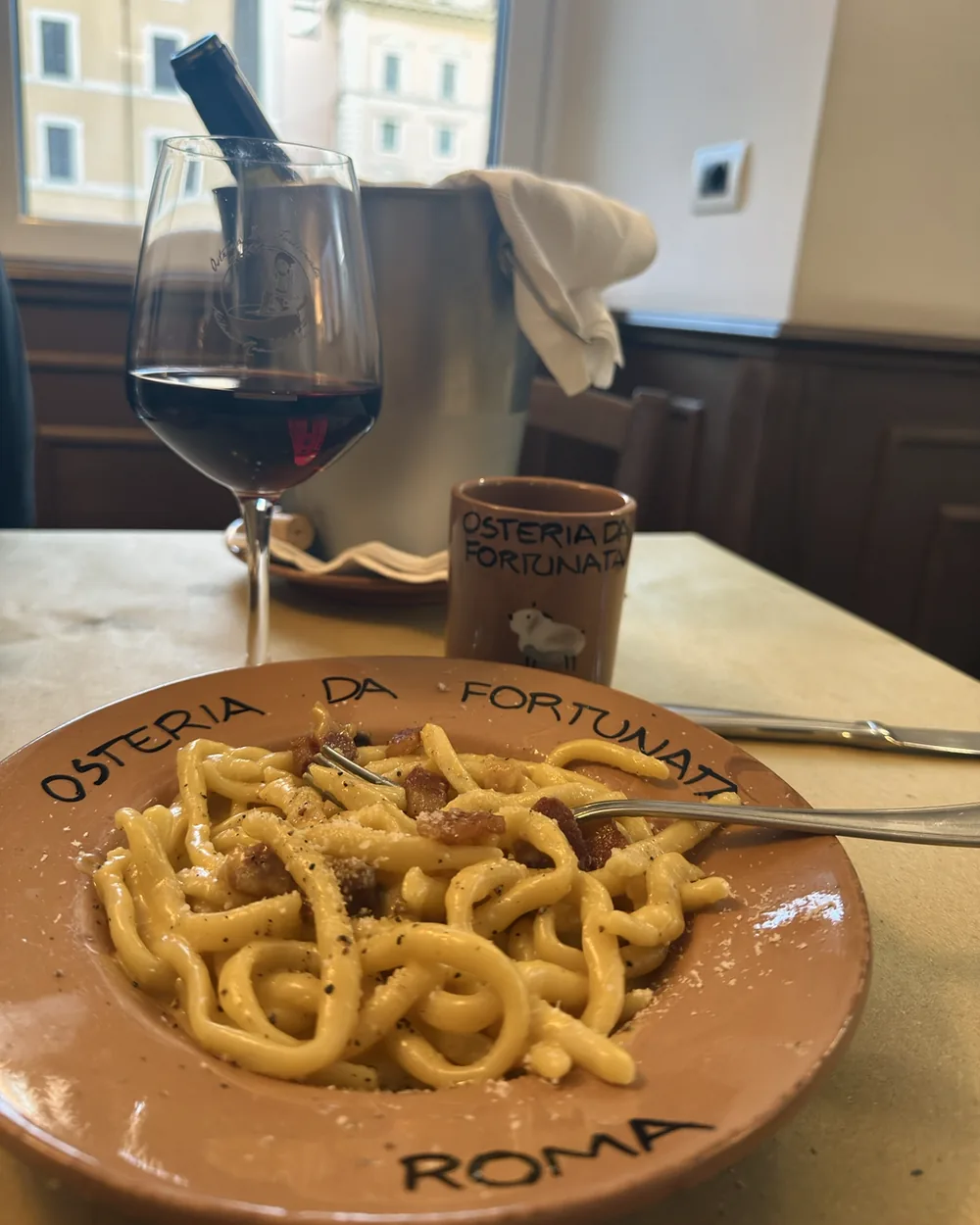 A terracotta plate of handmade carbonara with guanciale and black pepper, a glass of red wine and the piazza through the window at Osteria da Fortunata