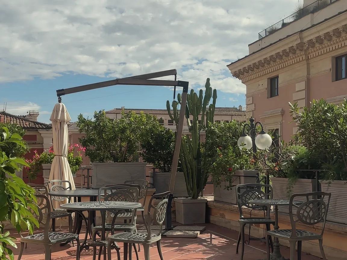 The rooftop garden terrace at Parlamento Boutique Hotel in Rome, with terracotta tiles, wrought-iron tables, potted greenery and a tall cactus, and a pink palazzo under a cloudy blue sky