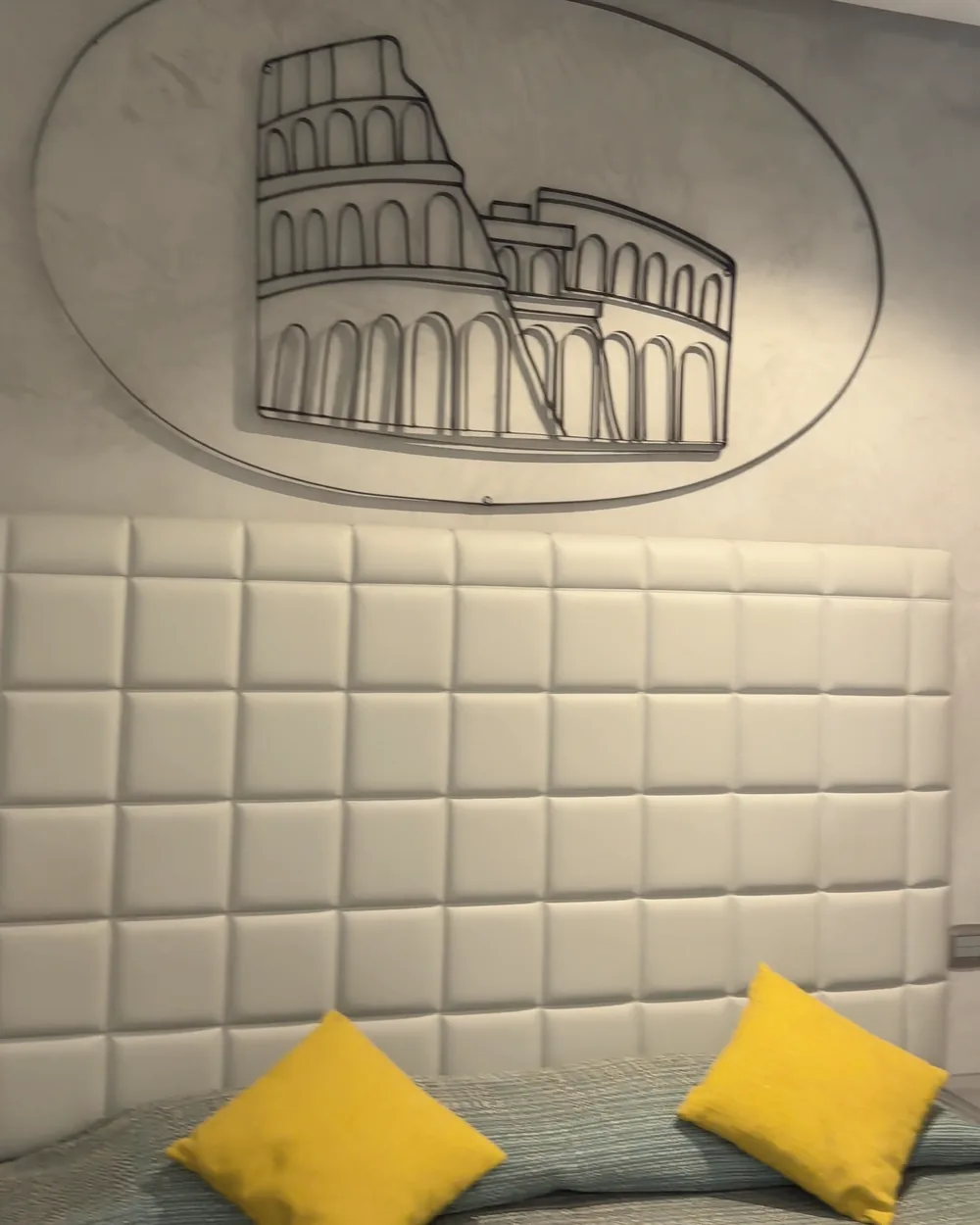 A white padded headboard with yellow cushions and a wrought-iron Colosseum outline on the grey plaster wall above it