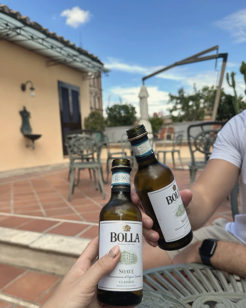 Two hands clinking small bottles of Bolla Soave Classico on the rooftop terrace