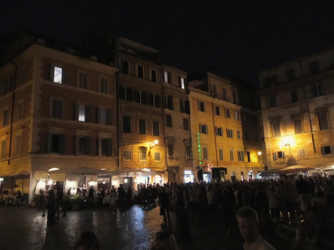 Golden lamplight on the ochre buildings and crowded cafe tables of Piazza di Santa Maria in Trastevere at night