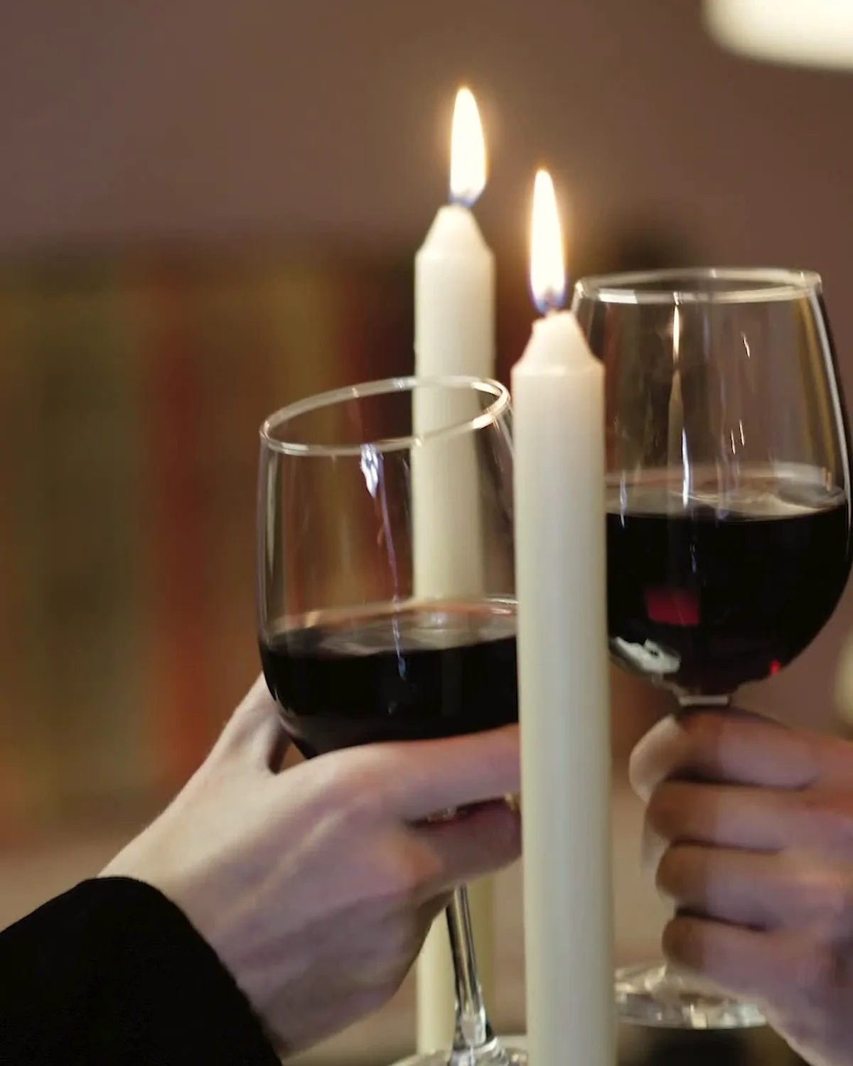 Two hands clinking glasses of red wine between tall white candles