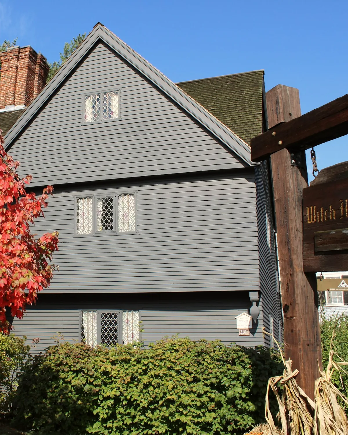 The dark grey, steep-gabled Witch House in Salem with diamond-pane windows and a red autumn tree