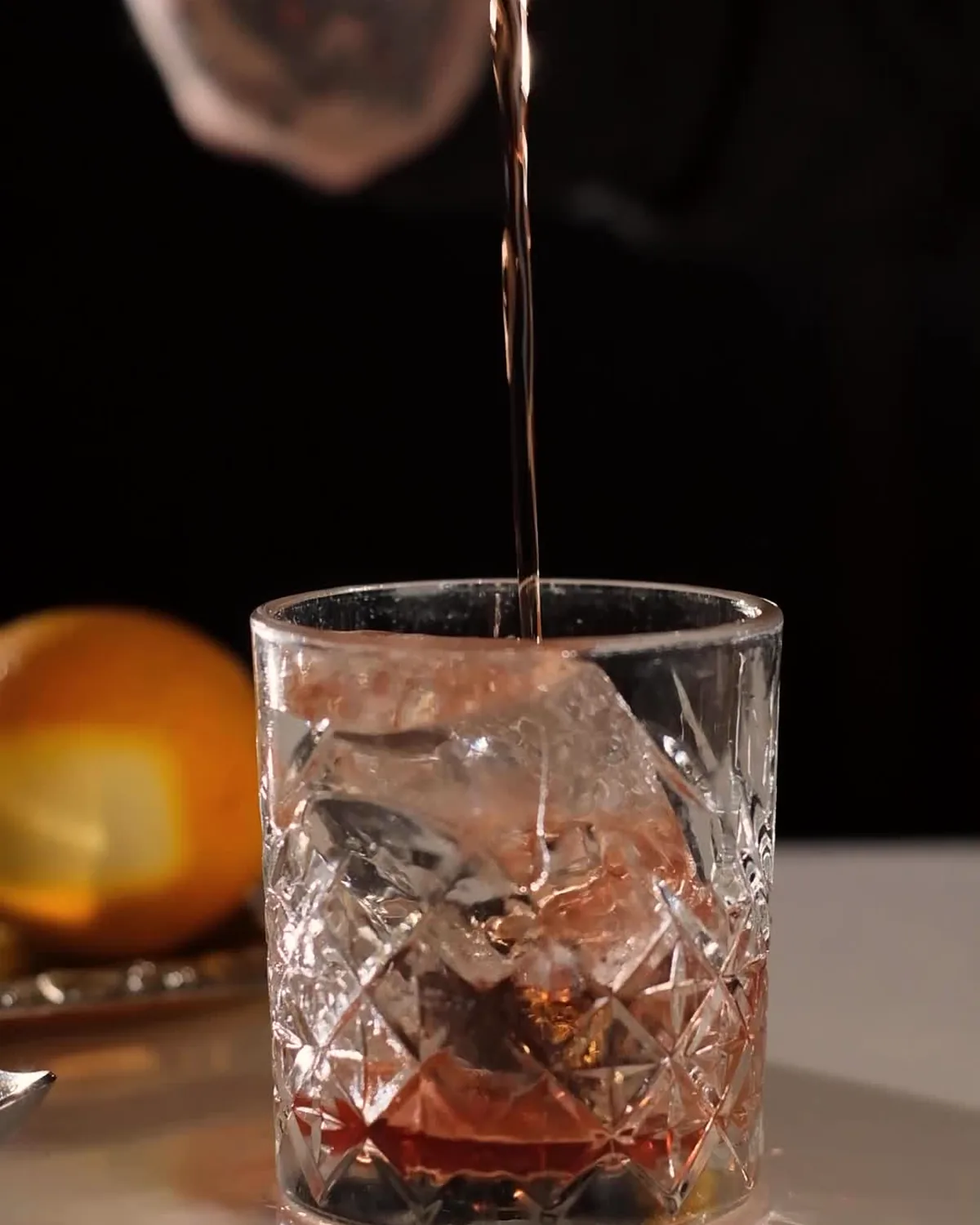 A dark cocktail being poured over ice into a cut-crystal glass beside an orange