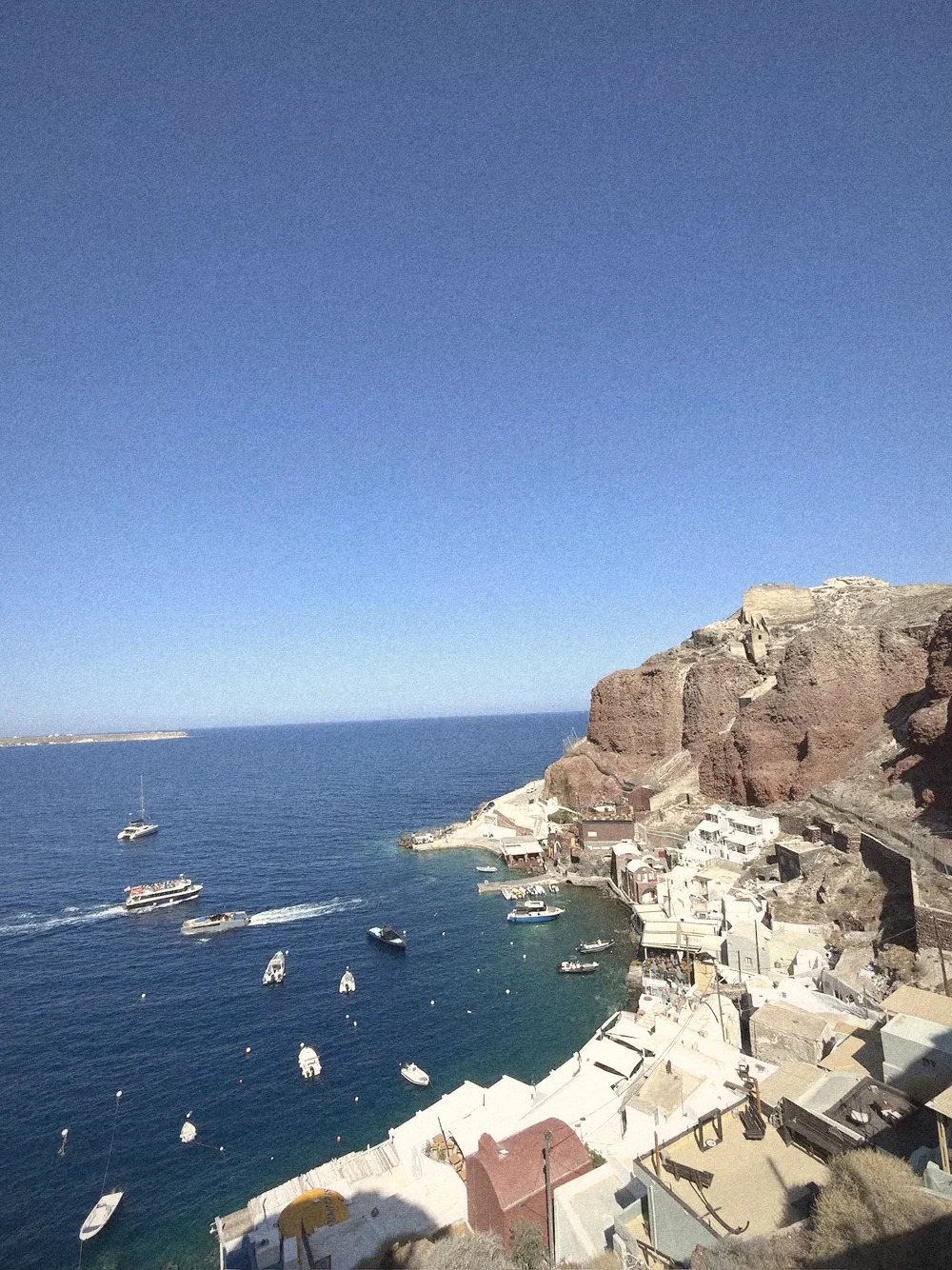 Amoudi Bay from above: deep blue water, red cliffs and white waterfront buildings with small boats