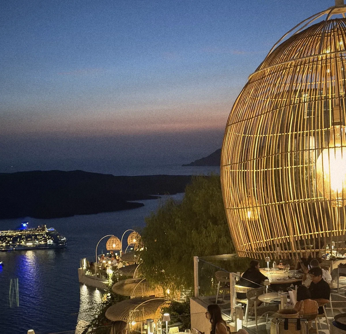 Barolo's terrace at dusk: a giant woven lantern glowing beside the caldera, with a lit cruise ship on the water below