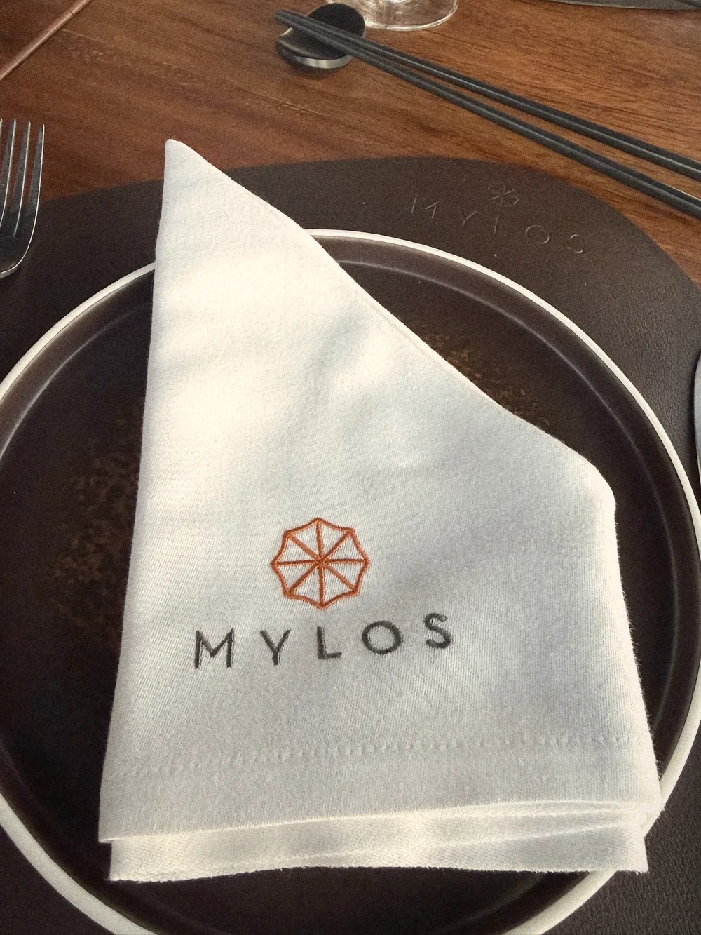 A white linen napkin embroidered with the orange Mylos windmill logo on a dark plate