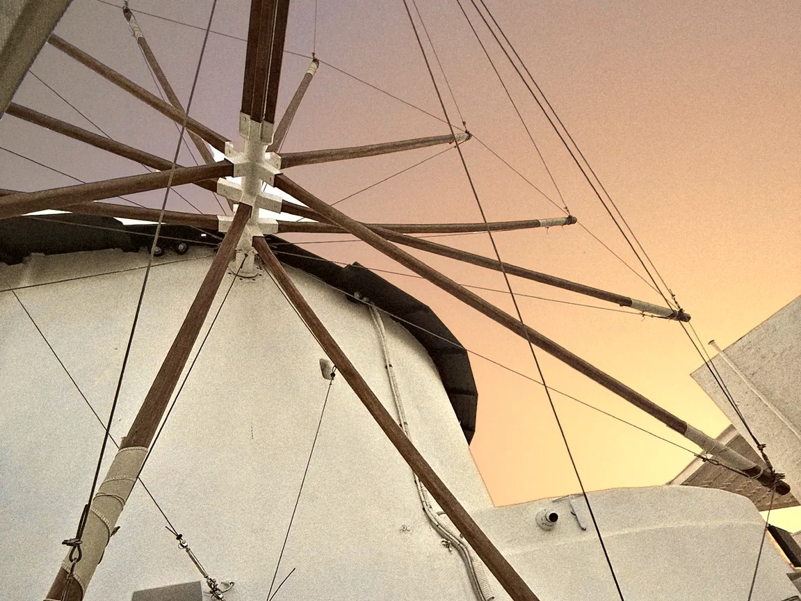 Looking up at the white Mylos windmill and its wooden sails against a peach and golden-orange evening sky in Firostefani, Santorini