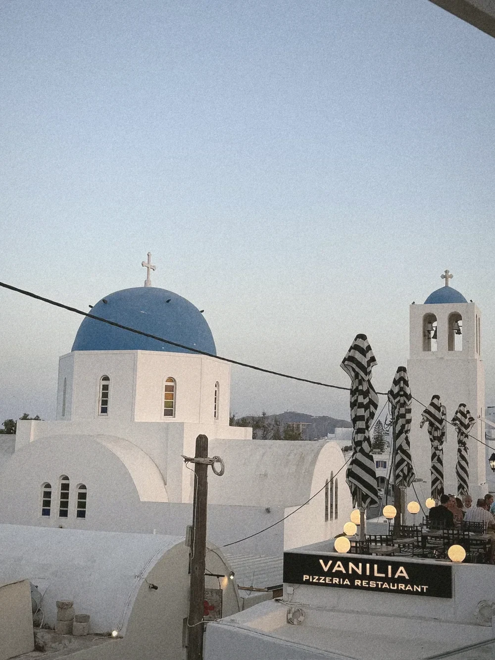 A whitewashed church with a blue dome and bell tower in Oia under a soft evening sky